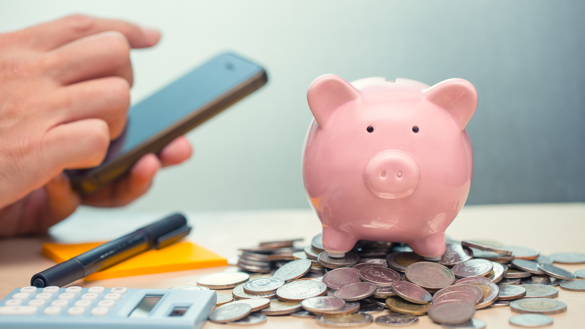 a person types on smartphone next to a piggy bank, which sits on top of a pile of coins, to represent a budget