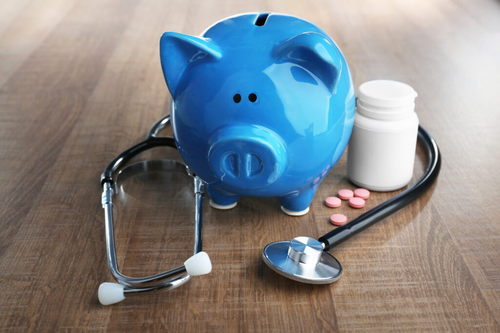 Stethoscope surrounds blue piggy bank and open bottle of pink pills | How to get a medical loan