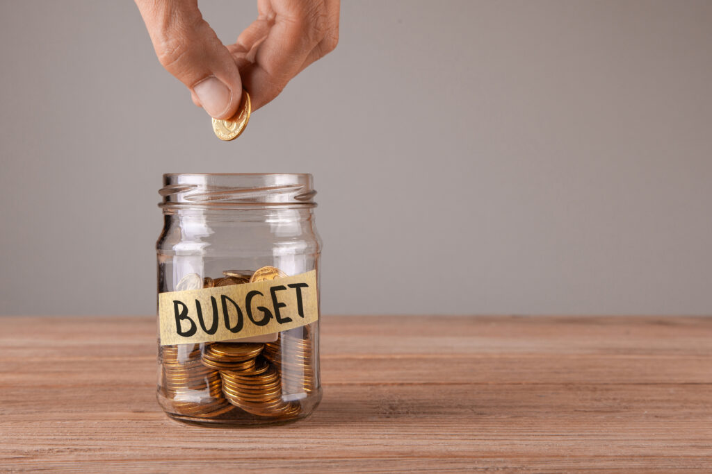 Close up of hand placing gold coin in jar labeled as budget | Planning for unexpected expenses