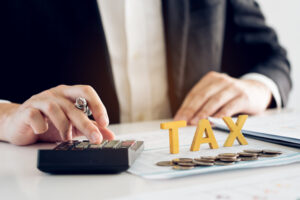 Person in a suit uses a calculator beside stacked coins and the word "TAX"