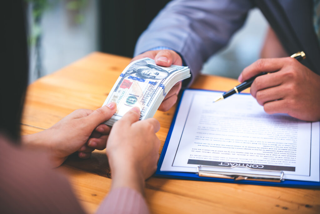 Hands exchanging a stack of hundred-dollar bills over a signed contract on a clipboard.